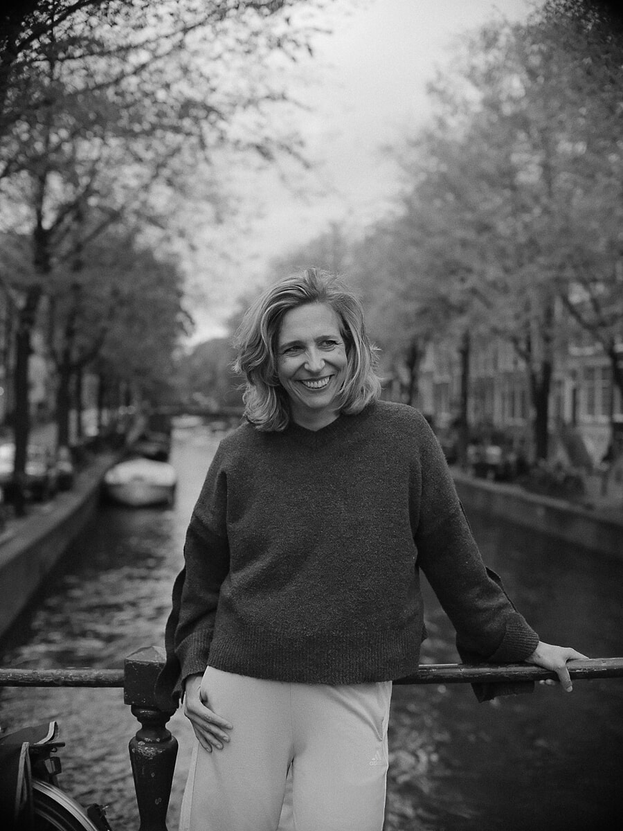 Blonde vrouw lacht terwijl ze achterover leunt tegen de balustrade langs de gracht, een zwart-wit confidenceportret tussen herfstbomen