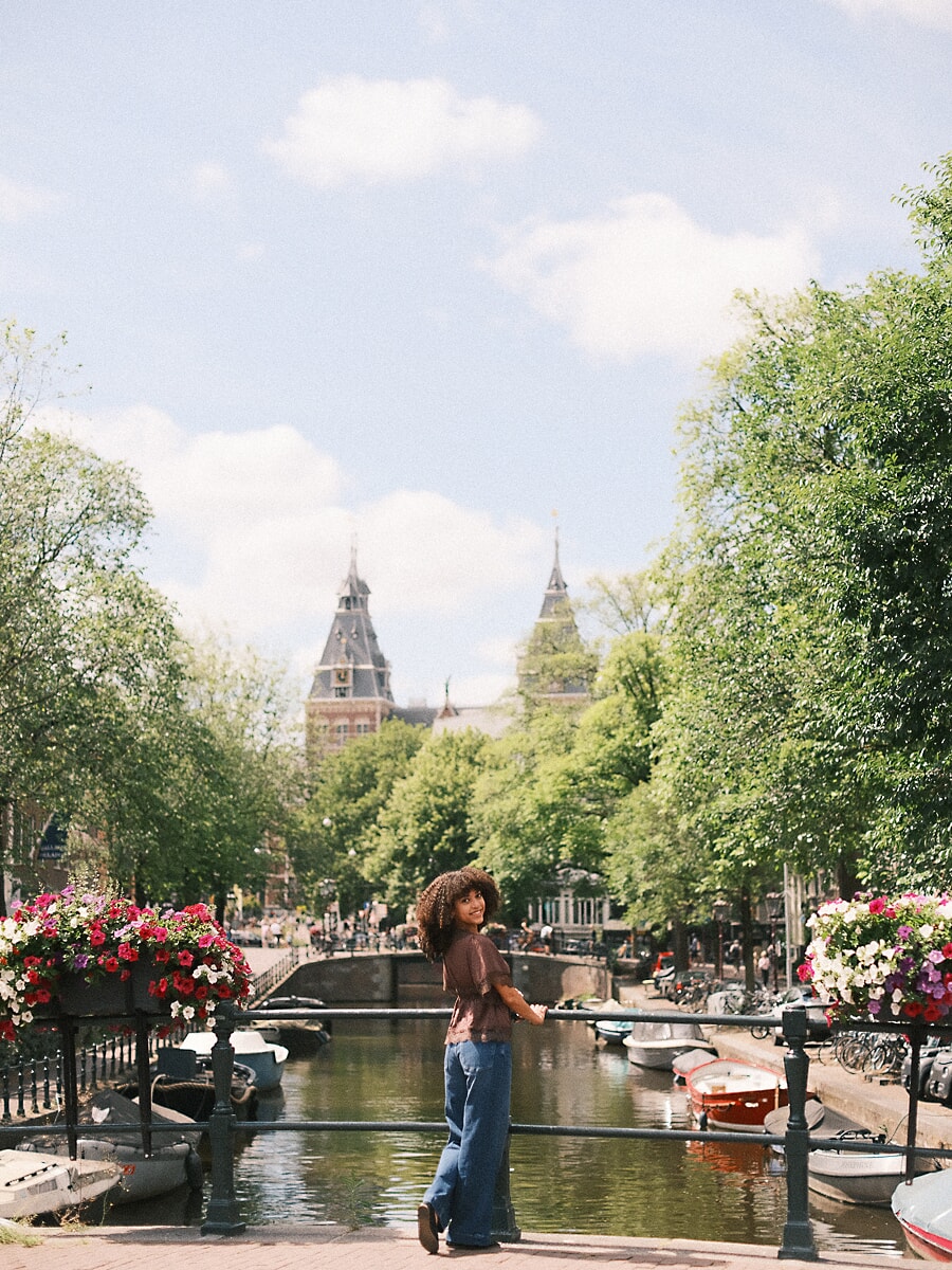 Vrouw met krullend haar kijkt om vanaf een brugleuning met de torens van het Rijksmuseum erachter, portretfotografie in Amsterdam