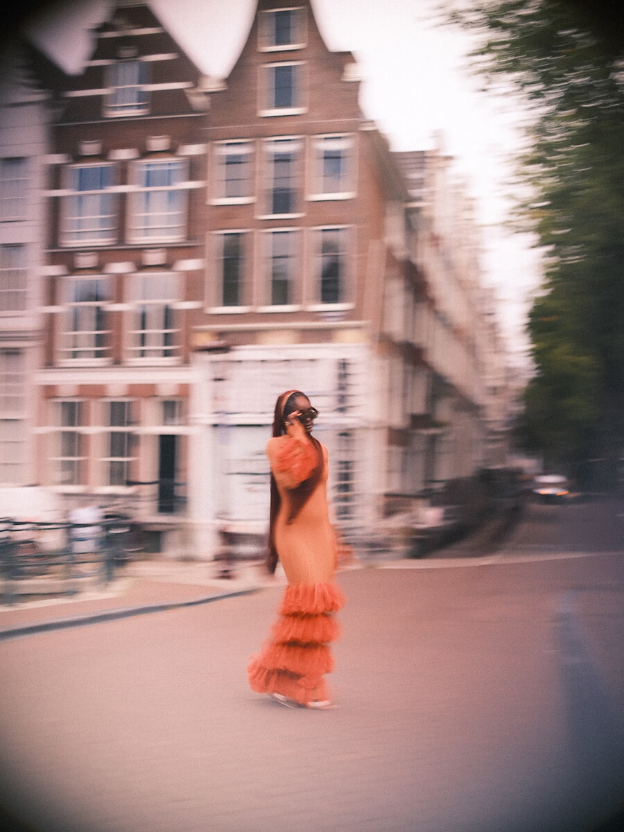 Vrouw in oranje jurk met ruches steekt de straat over in bewegingsonscherpte, met de gevels uitgesmeerd achter haar