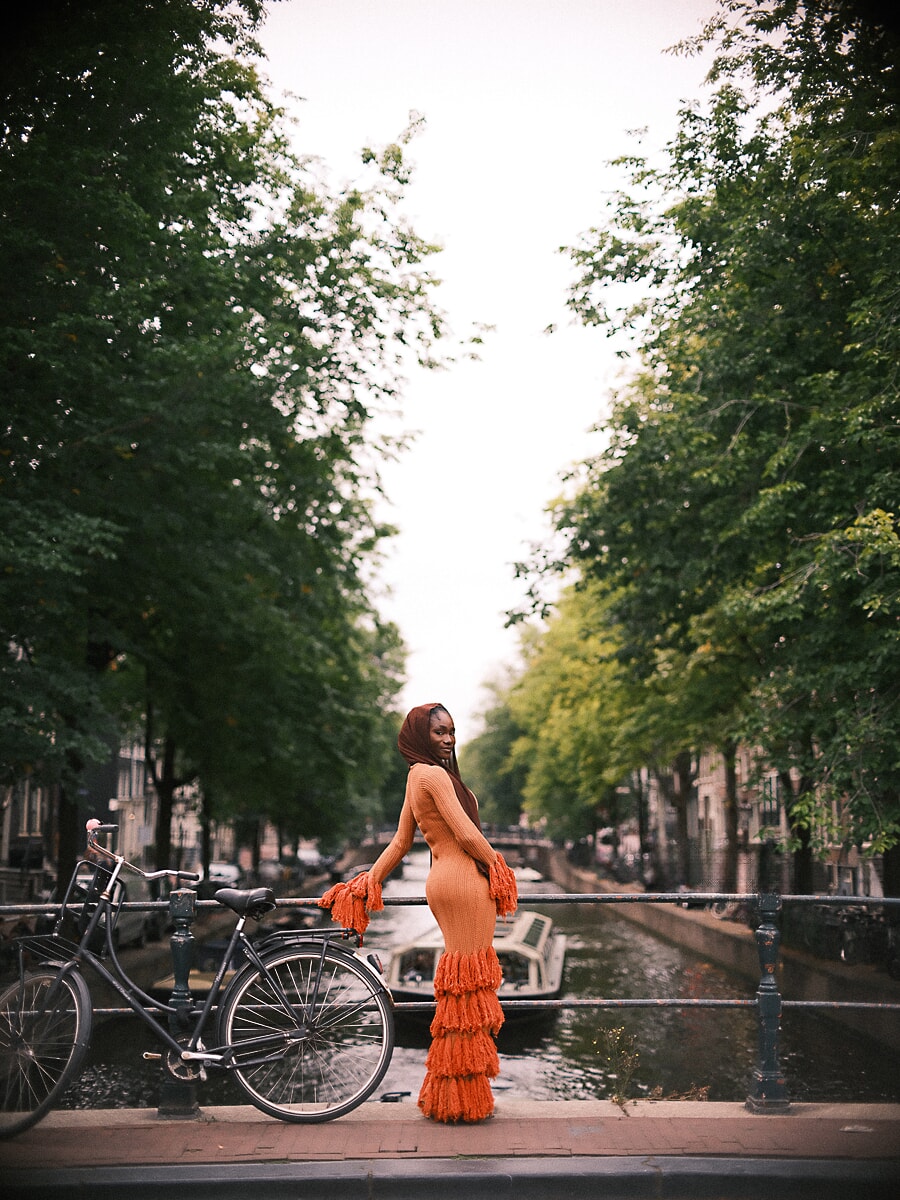 Vrouw in oranje gebreide jurk met franjes en hoofddoek kijkt om op een brug, een kleurrijk portret