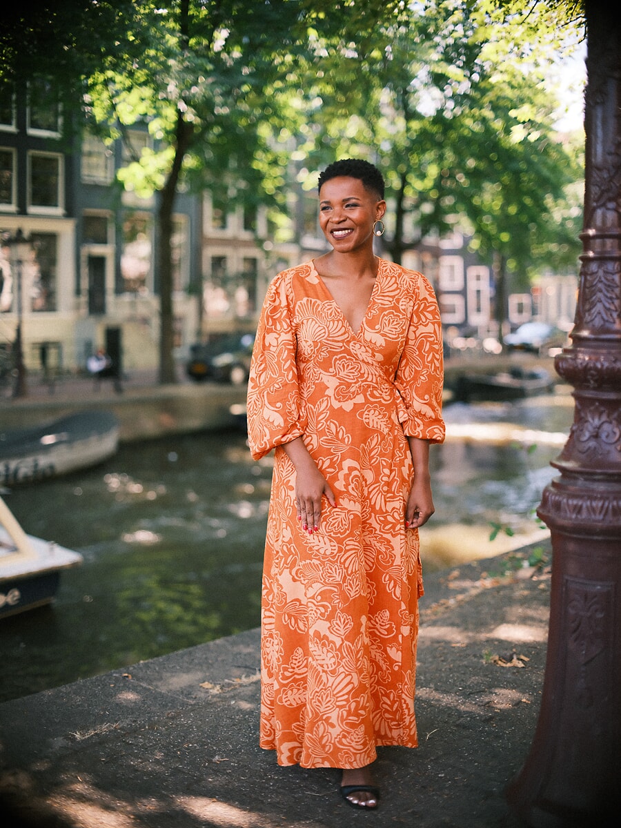Vrouw in oranje maxi-jurk met bladprint langs een Amsterdamse gracht met een sierlijke lantaarnpaal, een portret ten voeten uit
