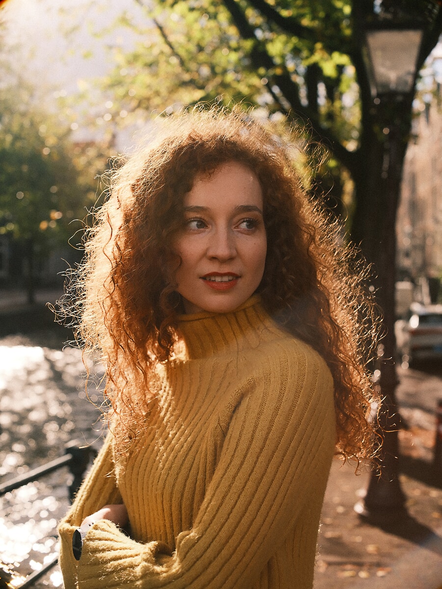 Vrouw met rode krullen in een okergele coltrui kijkt over haar schouder in gouden herfstlicht, portretfotografie in Amsterdam