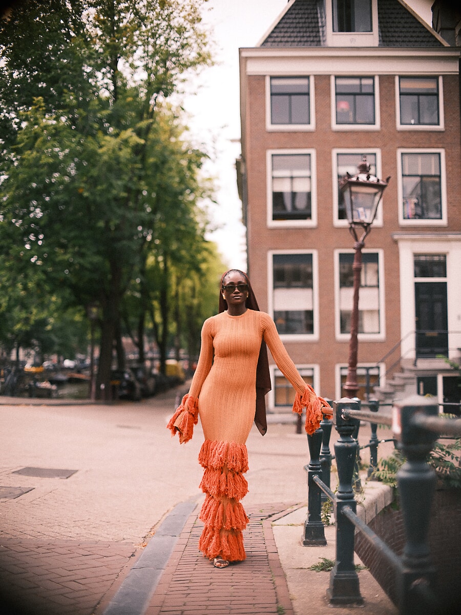 Vrouw in oranje jurk met franjes loopt over de kade onder een bakstenen trapgevel, een straatportret