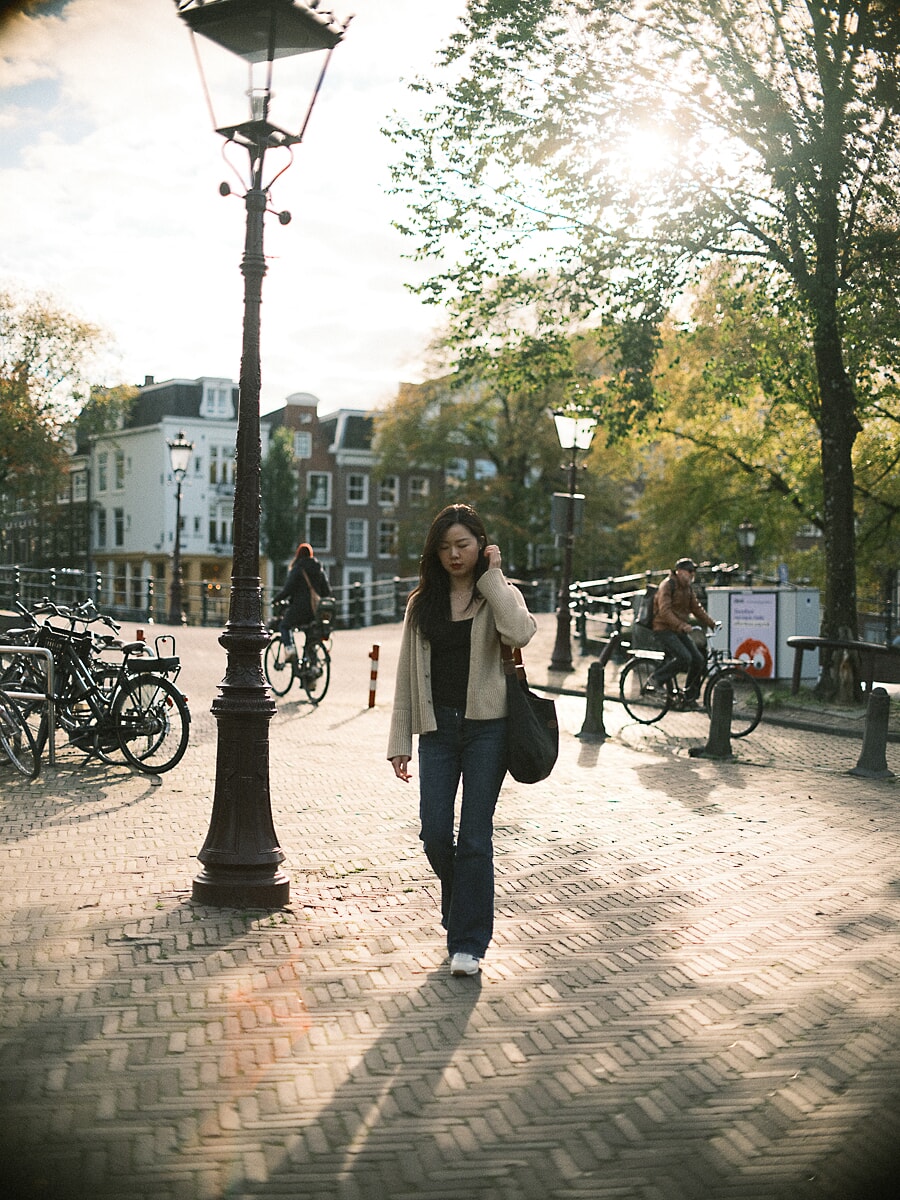Vrouw in crèmekleurig vest loopt over een bakstenen plein in laag herfstlicht, een ongedwongen datingportret in Amsterdam
