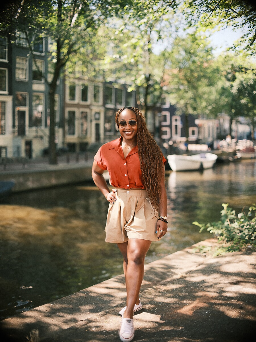 Vrouw in roestkleurige blouse en zonnebril wandelt langs een zonnige gracht tijdens haar portretfotoshoot in Amsterdam