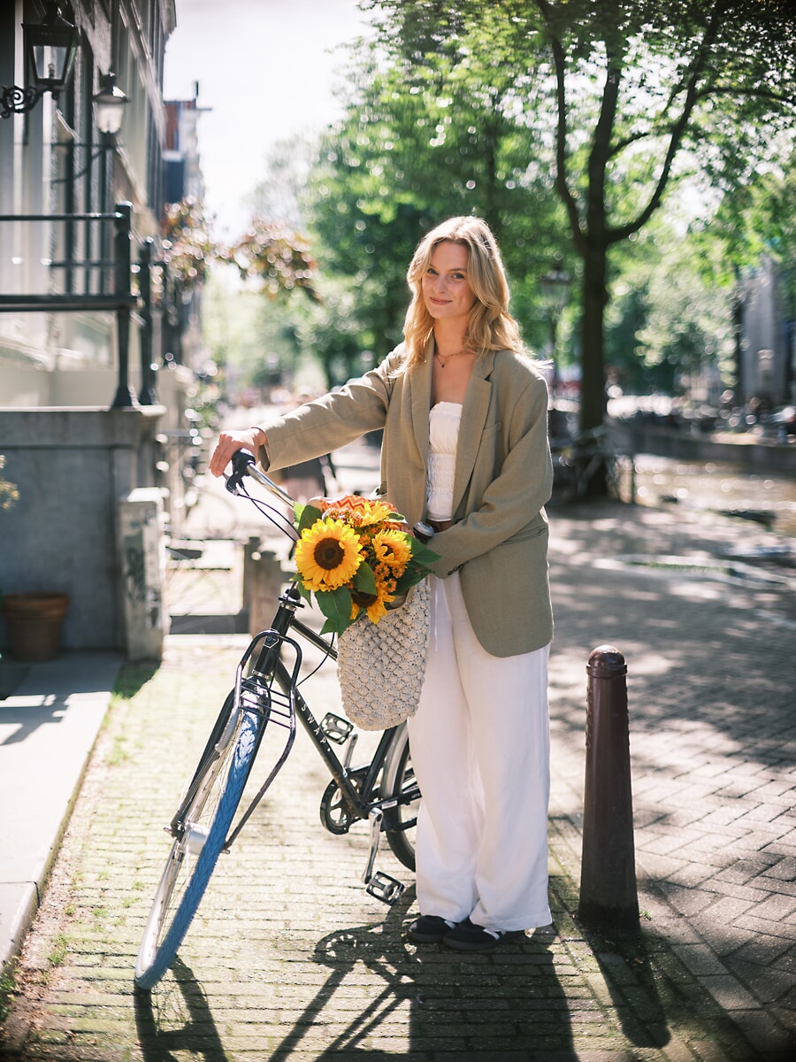 Vrouw in oversized blazer en witte broek bij een fiets met zonnebloemen in een netzak aan het stuur, een portret in Amsterdam