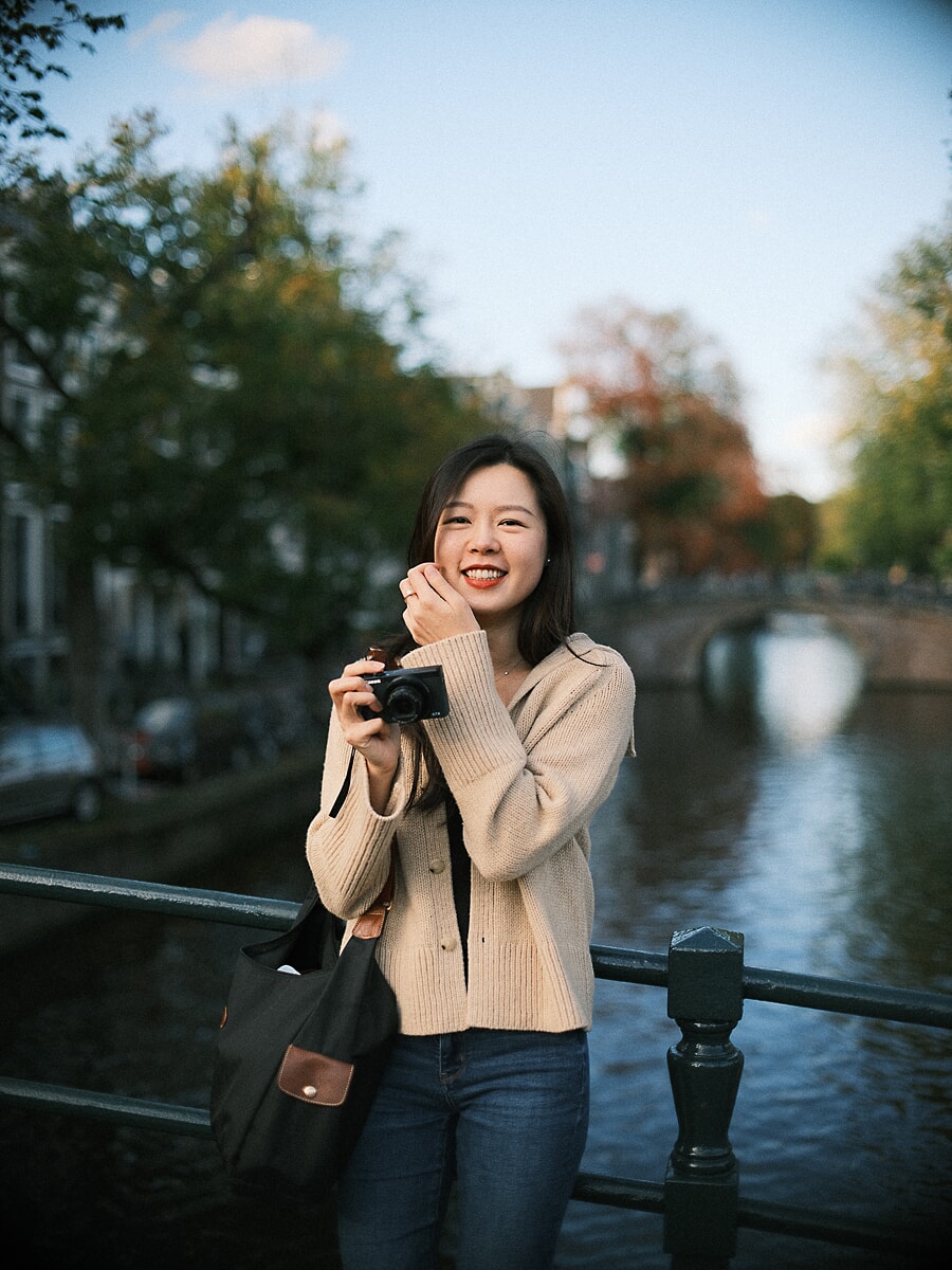 Vrouw in crèmekleurig vest lacht met een kleine camera in haar handen op een Amsterdamse grachtbrug, een spontaan beeld uit haar datingshoot