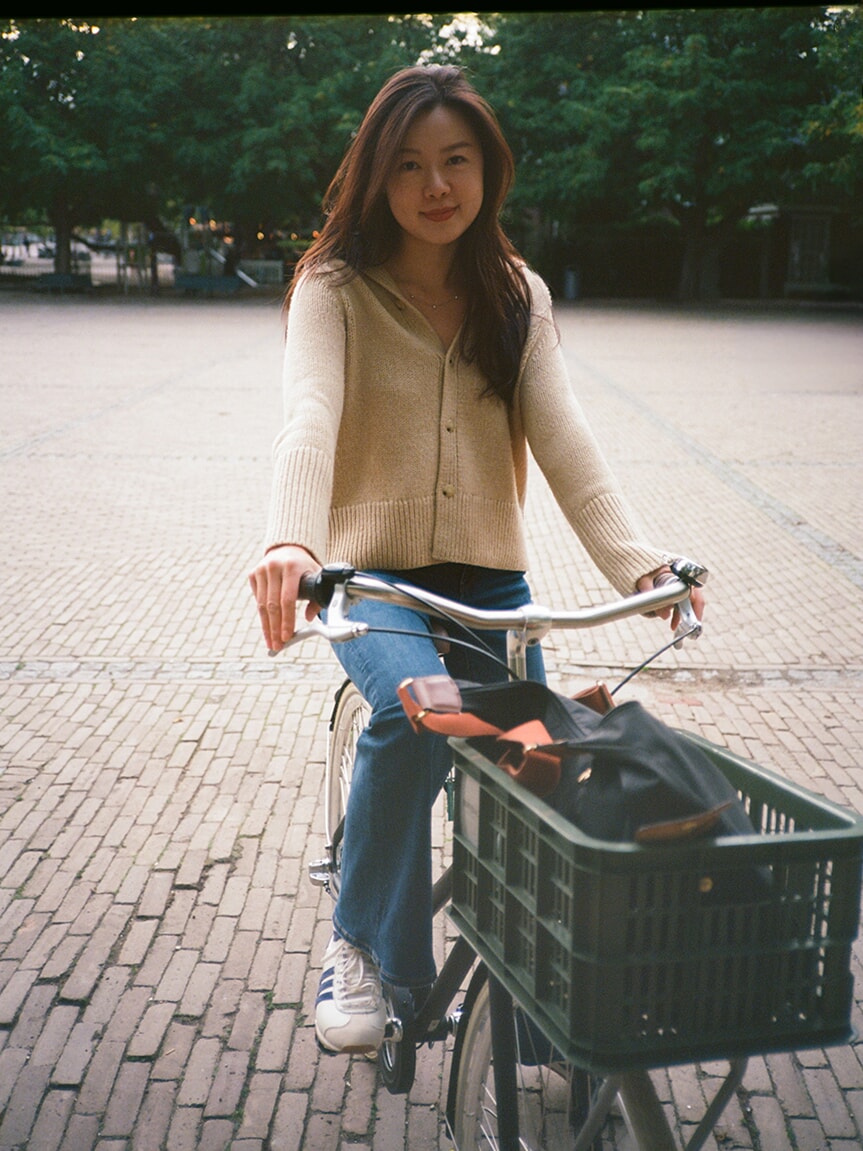 Portret van een vrouw in een crèmekleurig vest op een fiets met een zwart krat, tijdens een analoge fotoshoot in Amsterdam