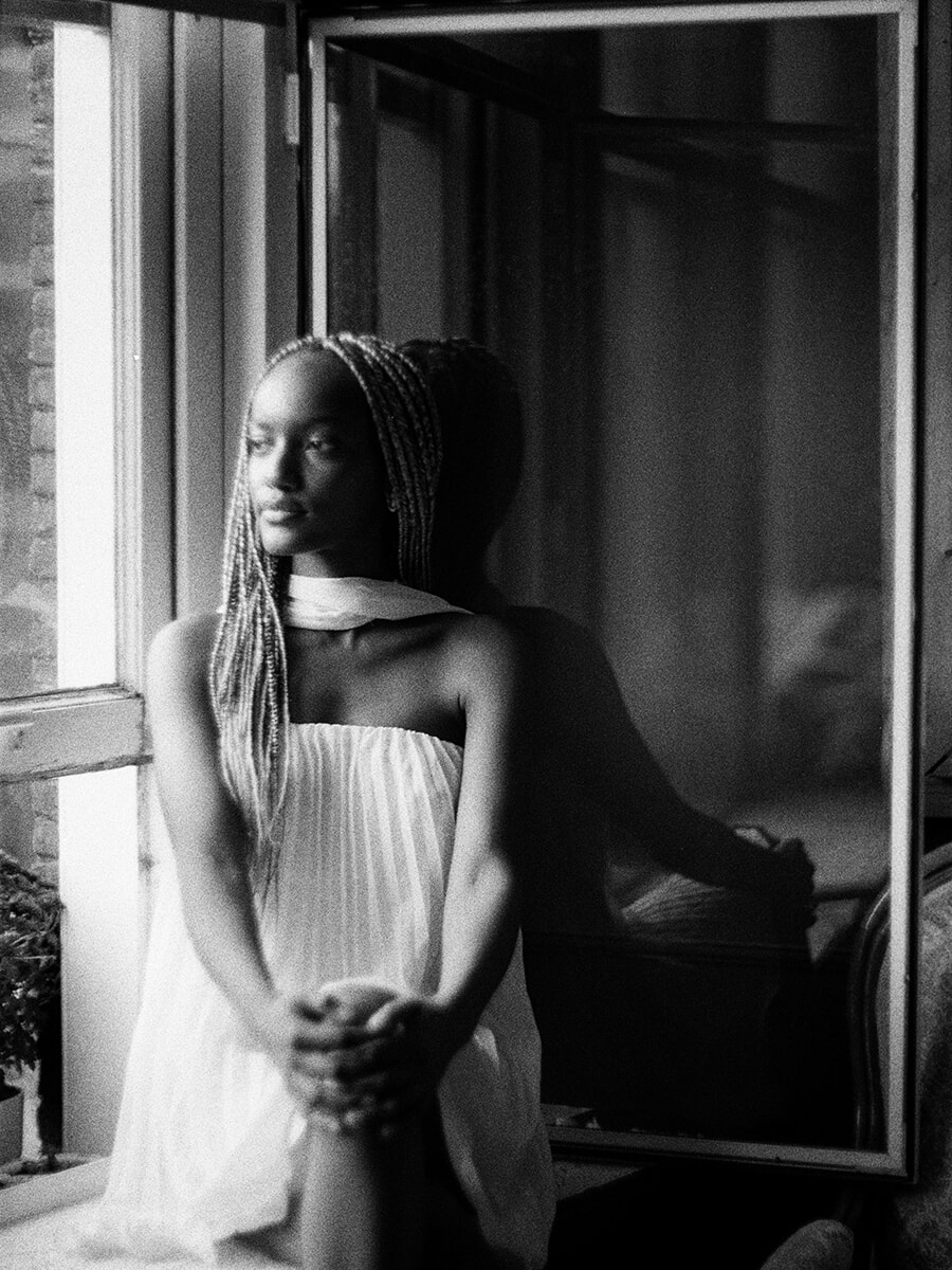 Bij een open raam zit een vrouw met lange vlechten en gevouwen handen, in een portret op zwart-witfilm