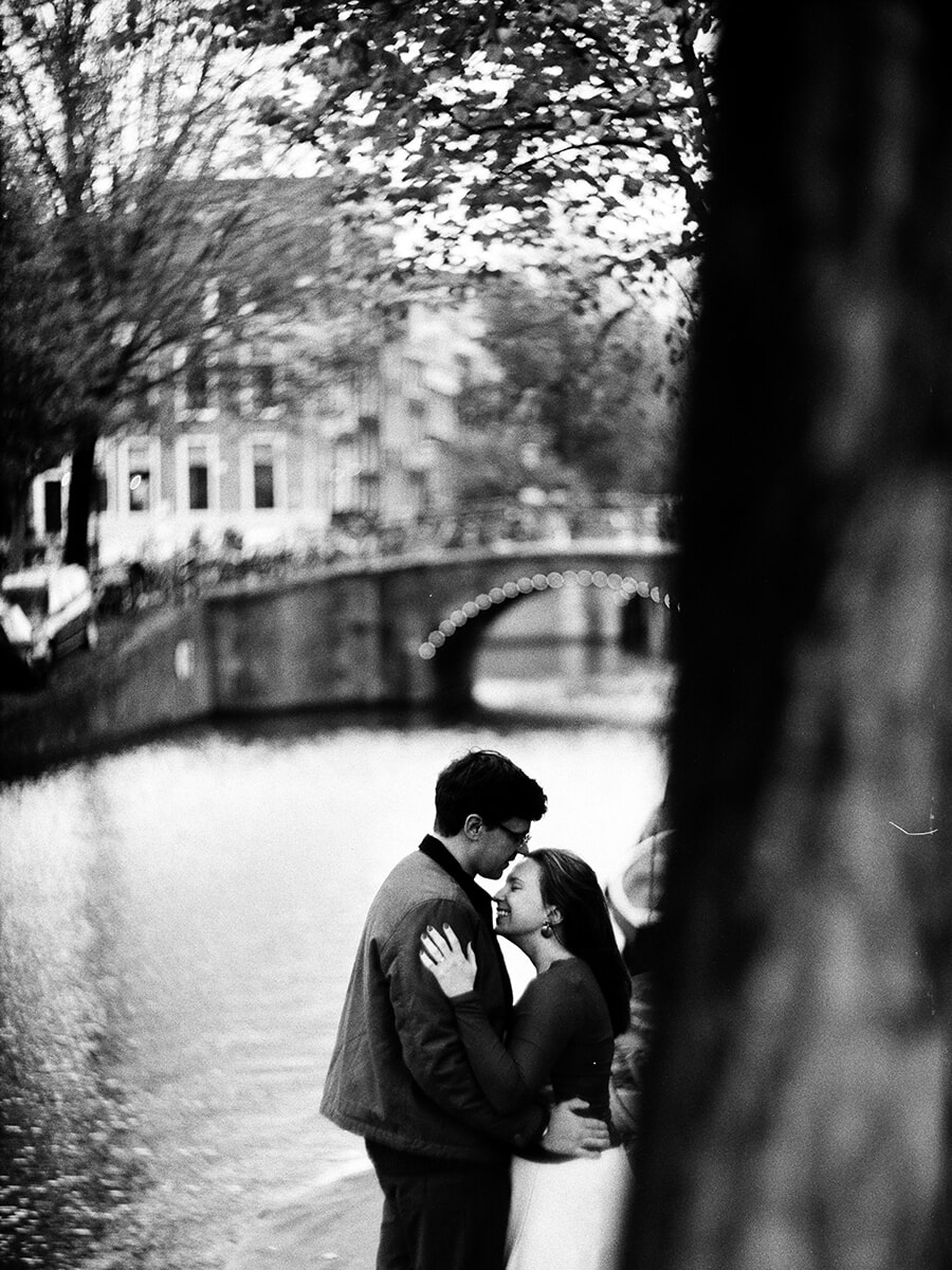 Een man kust zijn partner op het voorhoofd bij de gracht, met een onscherpe boomstam vooraan en een boogbrug op de achtergrond