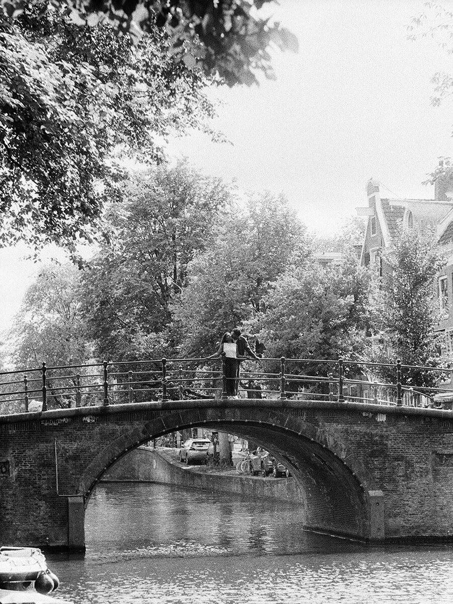 Een stel staat boven op een boogbrug tussen groene bomen, vastgelegd tijdens een analoge koppelfotoshoot in Amsterdam