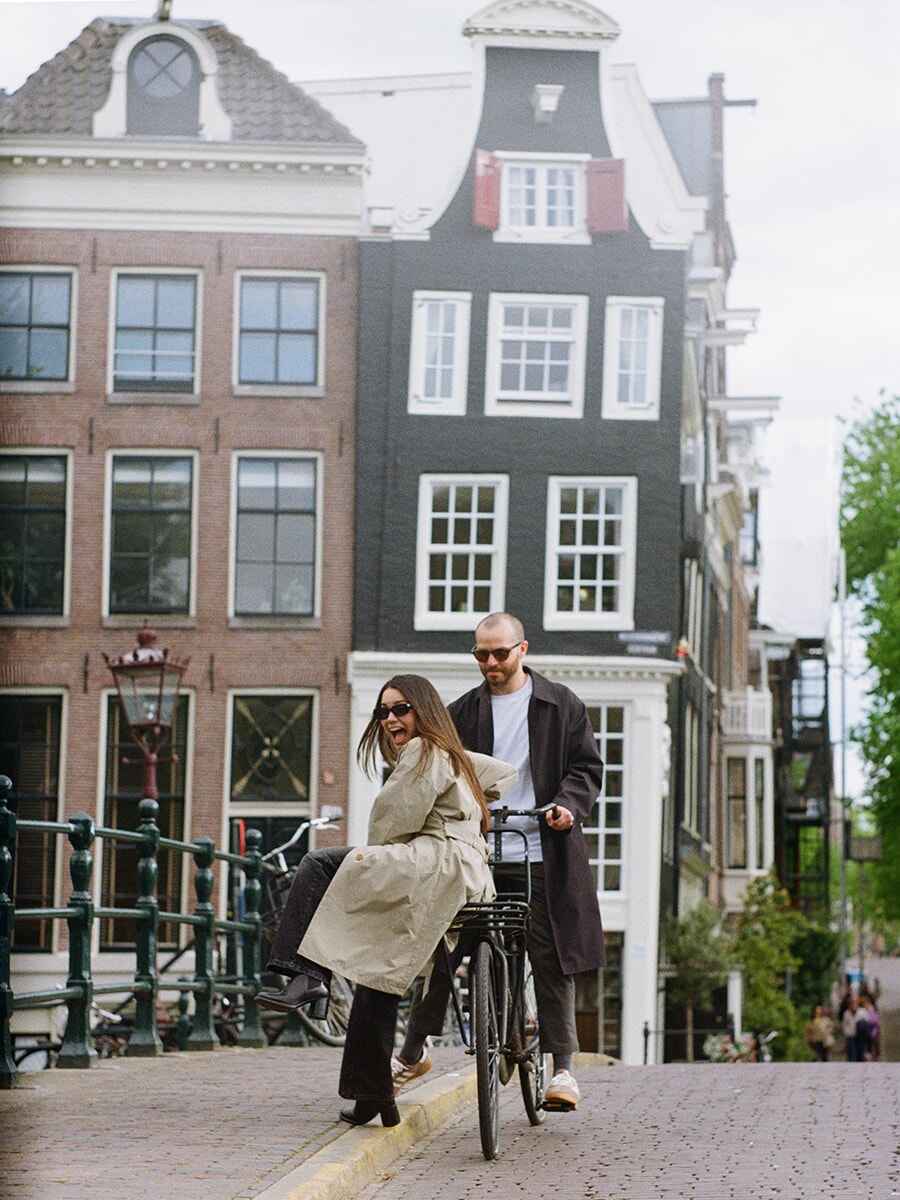 Een vrouw lacht op de voordrager van een fiets, haar partner erachter, tijdens een analoge koppelfotoshoot op een brug in Amsterdam