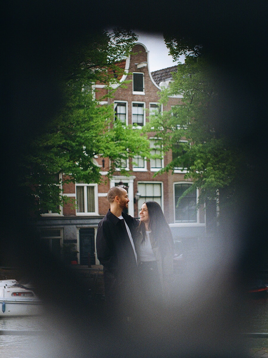 Een stel glimlacht naar elkaar aan een gracht in Amsterdam, gezien door een donkere, onscherpe voorgrond en vastgelegd op kleurenfilm