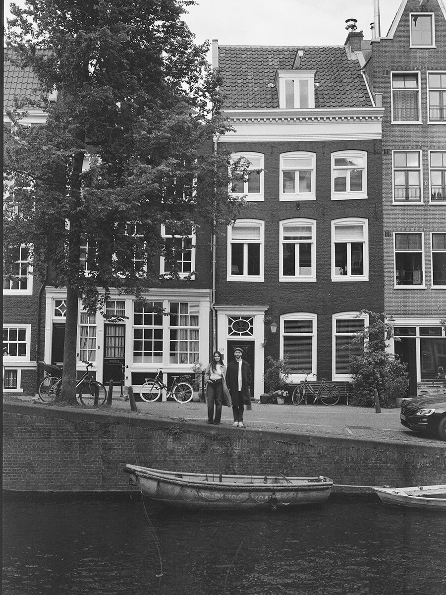 Analoge koppelfoto in Amsterdam, met het stel aan de overkant van de gracht, hoge bakstenen huizen erachter en een roeiboot ervoor