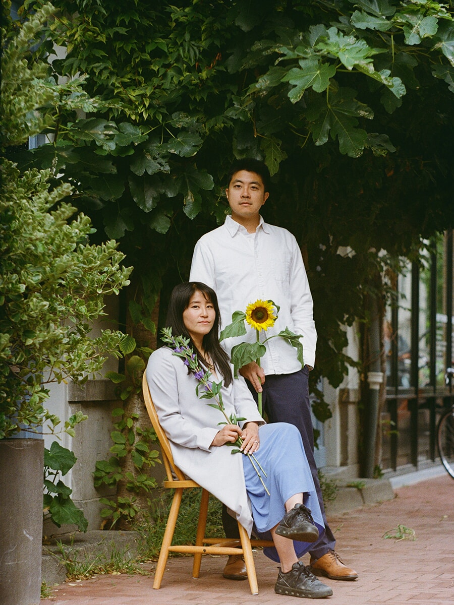 Tussen het groen zit een vrouw met paarse bloemen op een houten stoel, naast haar staande partner met een zonnebloem