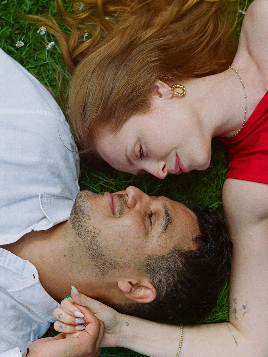 Van bovenaf gezien ligt een stel hand in hand in het gras tijdens een analoge fotoshoot in Amsterdam, zij in rood en hij in wit