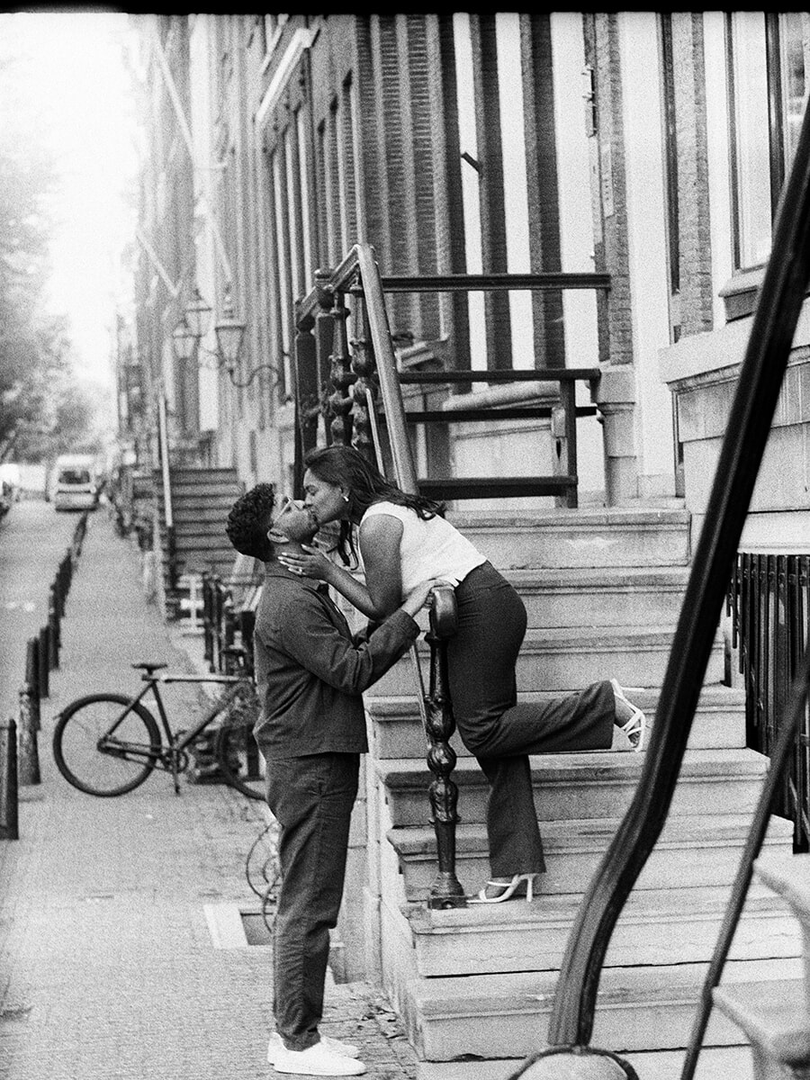 Vanaf een stenen trap in Amsterdam kust een vrouw haar partner met één been naar achteren gebogen, tijdens een analoge koppelfotoshoot