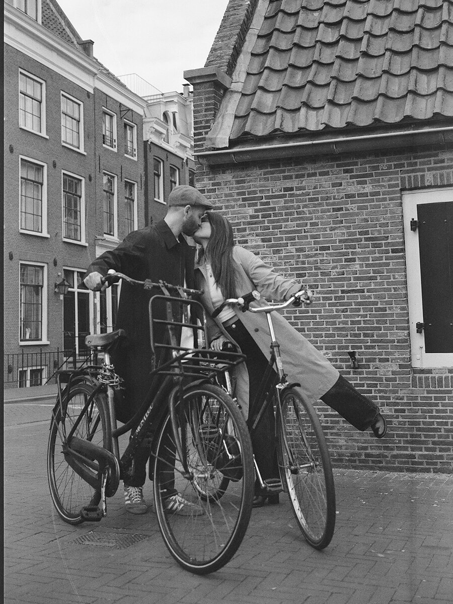 Naast twee fietsen bij een bakstenen muur in Amsterdam kust een stel elkaar op zwart-witfilm, terwijl zij één been naar achteren optilt