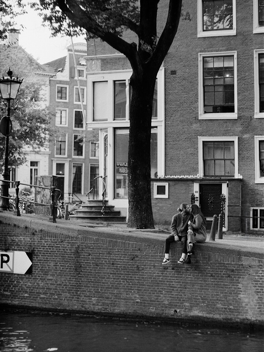 Een kussend stel zit onder een boom op een kademuur in Amsterdam, met grachtenpanden erachter, vastgelegd op zwart-witfilm