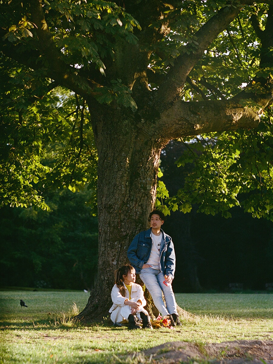 Analoge koppelfotoshoot in een park in Amsterdam, met een vrouw aan de voet van een grote boom en haar partner tegen de stam