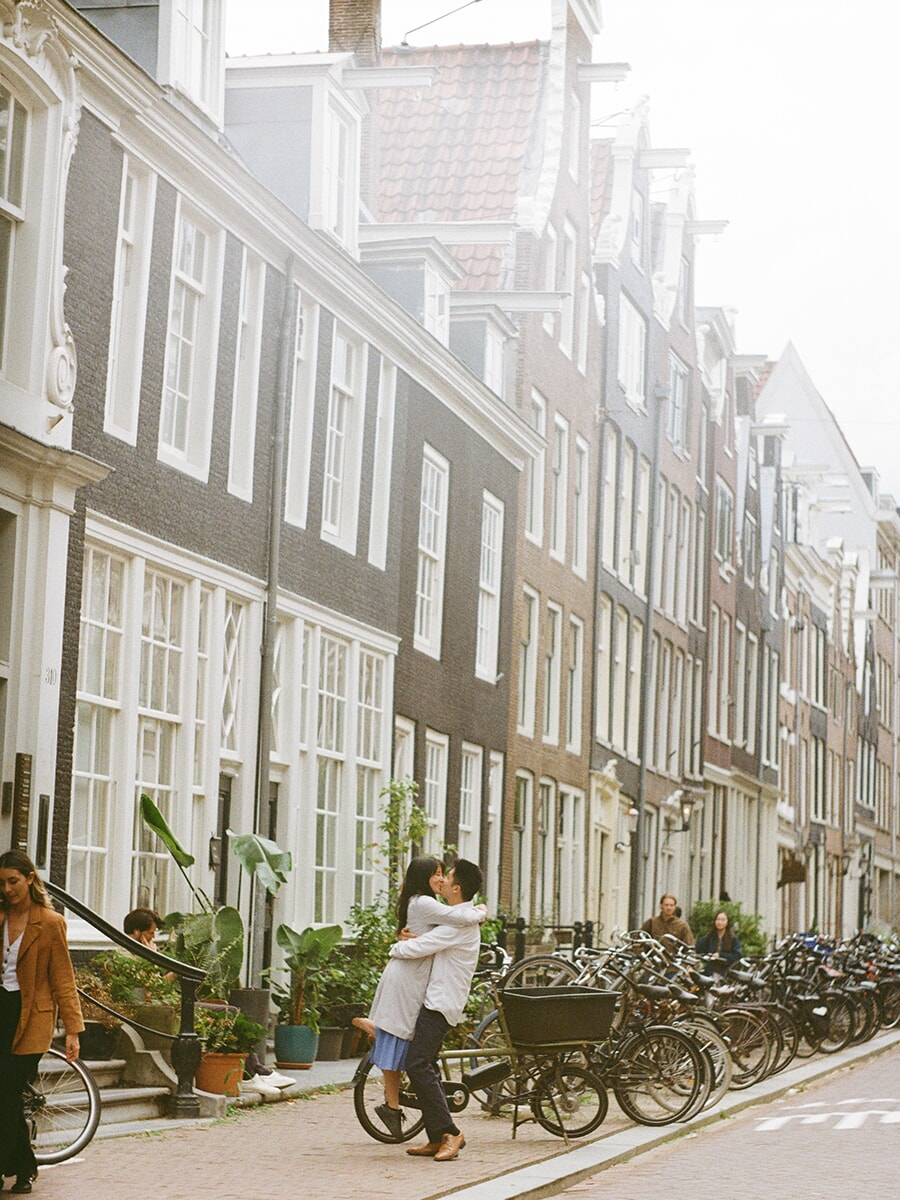 Tijdens een analoge loveshoot in Amsterdam tilt een man zijn partner op voor een kus, naast fietsen en hoge grachtenpanden