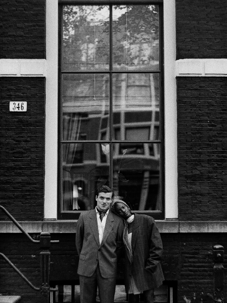 Onder een hoog raam met weerspiegelde gevels legt een vrouw haar hoofd op de schouder van haar partner