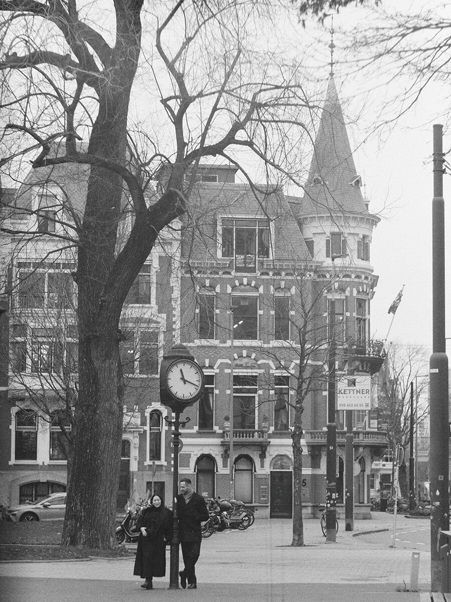 Een stel onder een straatklok, met een hoog bakstenen gebouw met torentjes erachter, in een ruim opgezet portret op zwart-witfilm