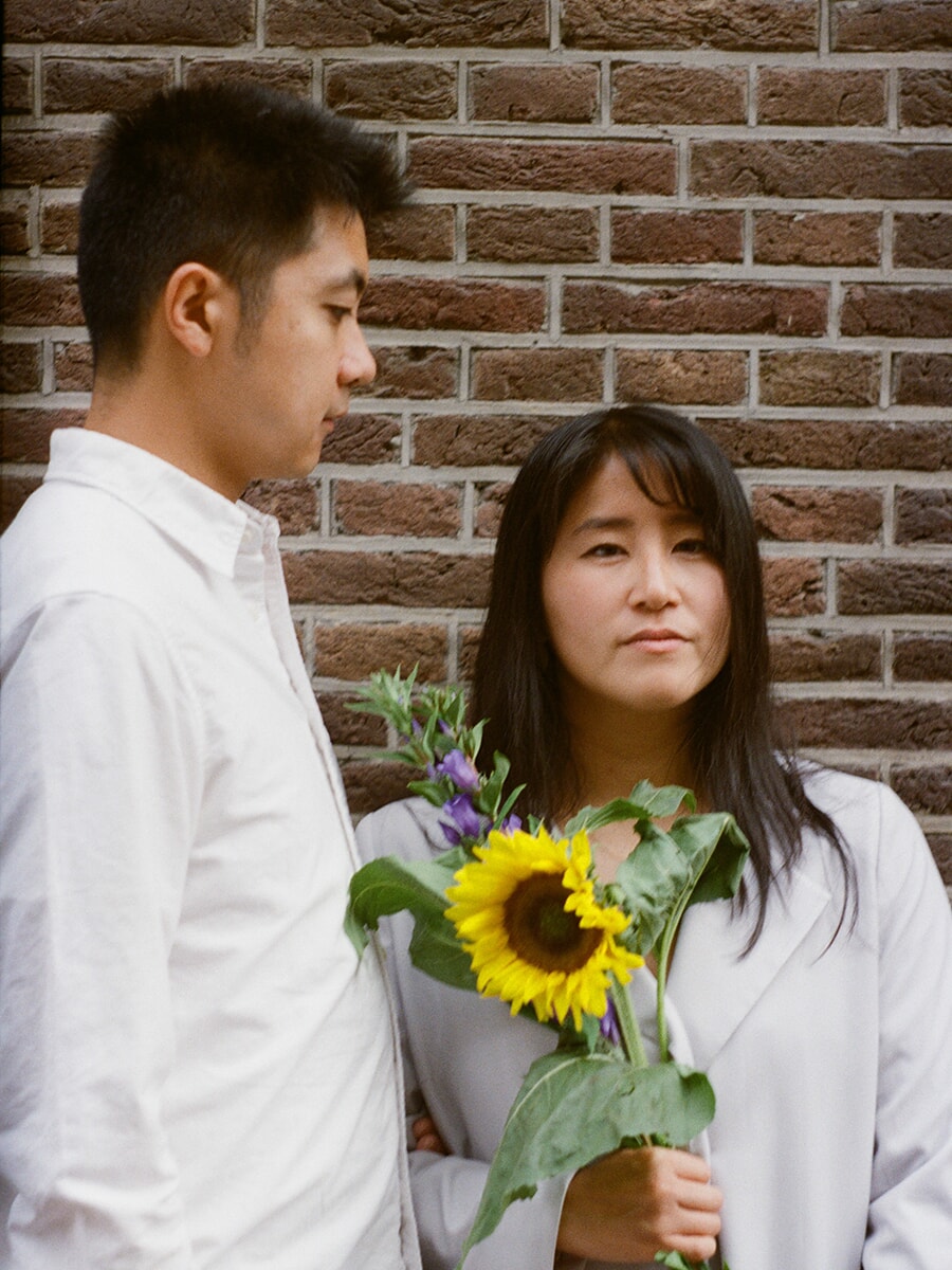 Koppelportret op kleurenfilm tegen een bakstenen muur, met een vrouw die een zonnebloem en paarse bloemen vasthoudt naast haar partner