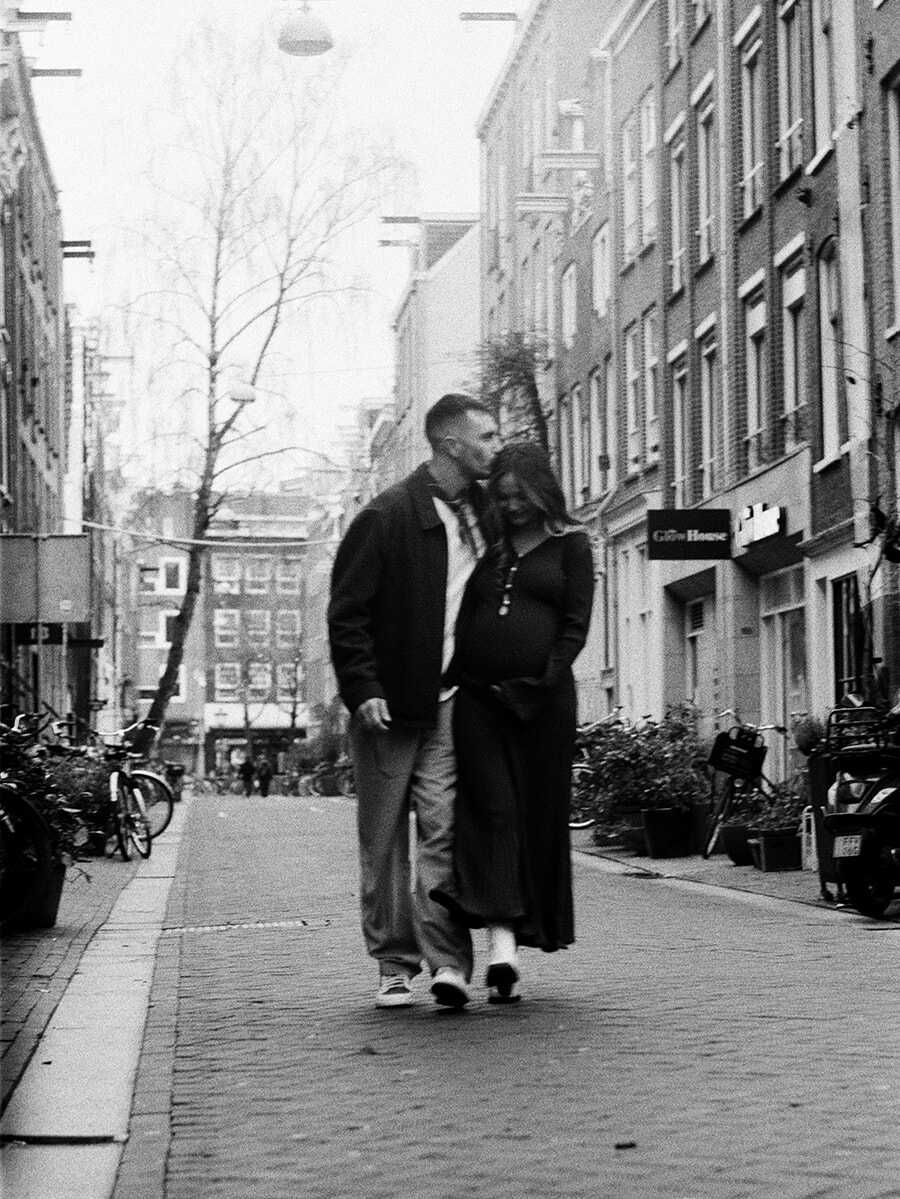 Tijdens een zwangerschapsfotoshoot op zwart-witfilm lopen aanstaande ouders door een smalle straat in Amsterdam, terwijl hij haar op het hoofd kust