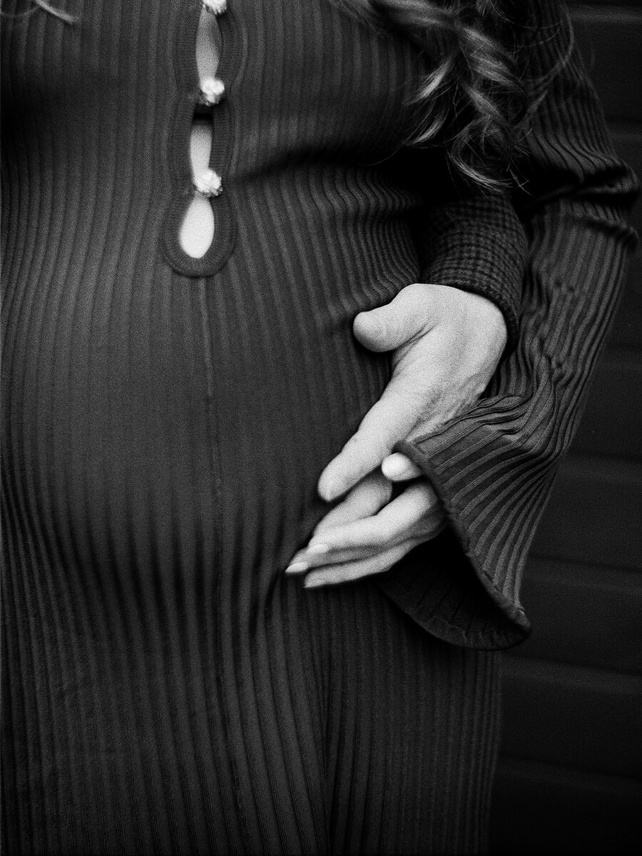 Detail van een zwangerschapsfotoshoot op zwart-witfilm, met handen samen op een zwangere buik in een donkere ribjurk