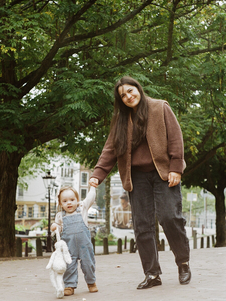 Moeder en peuter lopen hand in hand onder de bomen tijdens een analoge familiefotoshoot in Amsterdam, met een wit knuffelkonijn erbij