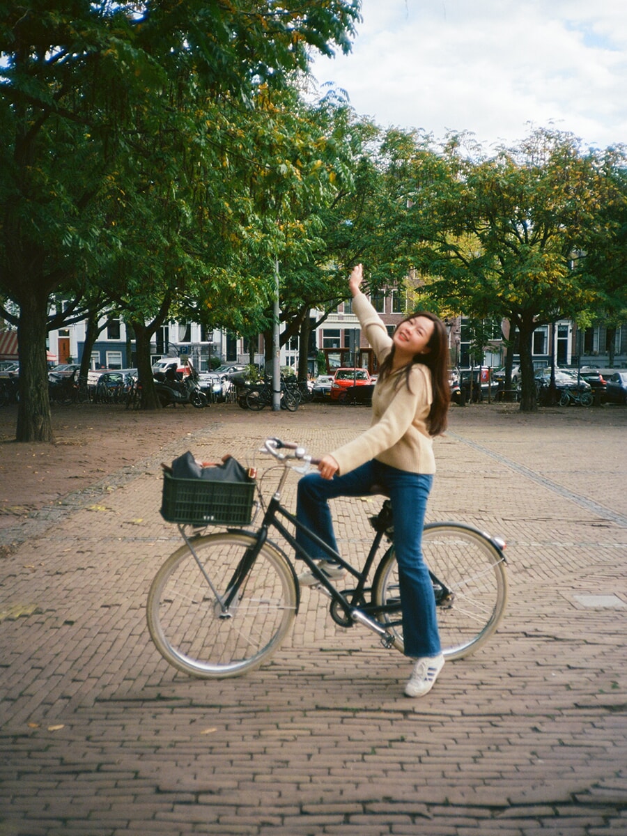 Op een plein met klinkers steekt een lachende vrouw in een crèmekleurig vest één arm omhoog terwijl ze op haar fiets zit